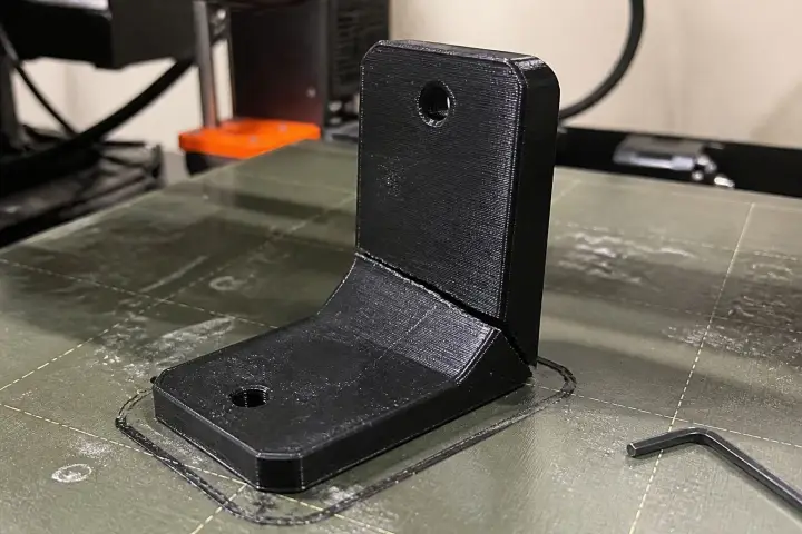 A black 3D printed bracket on a printer bed with an allen key beside it