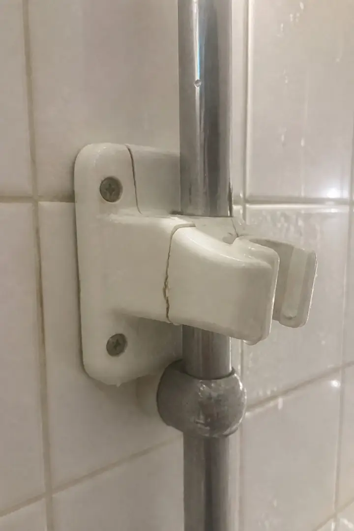 A cracked white shower head bracket clamped to a chrome rail on a tiled wall