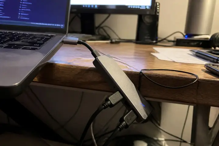 A USB hub hanging off a laptop port on a wooden desk, with cables trailing below it