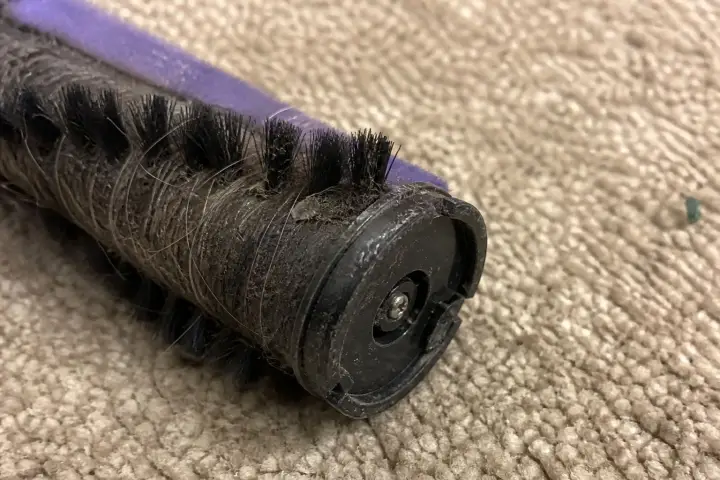 A vacuum brush roller wound with hair, lying on a carpet with its end cap facing the camera